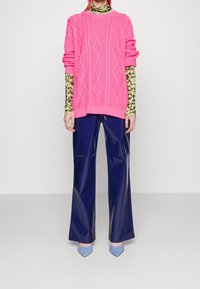 Rosa kabelstickad oversized tröja, lager på lager över en grön och svart mönstrad topp, med högblanka marinblå vida byxor och ljusbl å spetsiga skor.