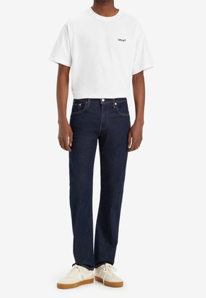 Camiseta blanca de algodón con el logo negro de Levi's, combinada con jeans de mezclilla azul oscuro y zapatillas blancas con suelas de color beige. Conjunto casual con líneas limpias.