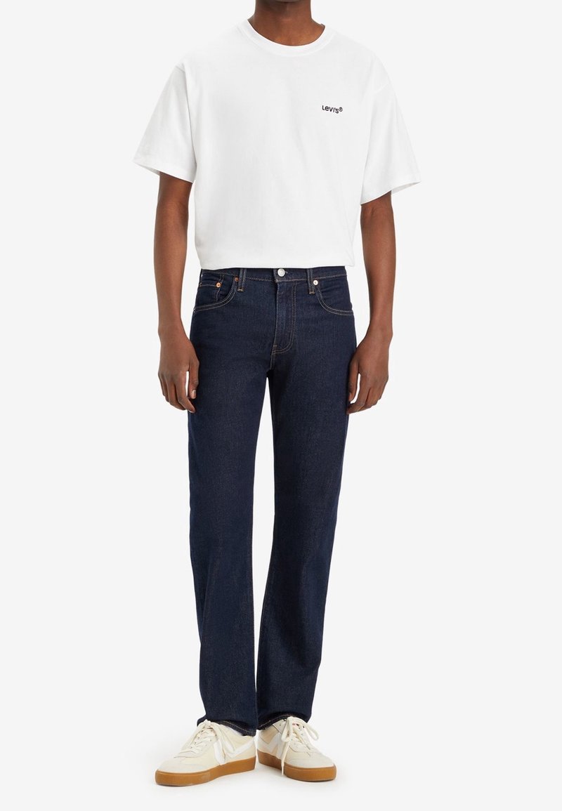 Vit bomulls t-shirt med svart Levi's-logotyp, tillsammans med mörkblå denimjeans och vita sneakers med tanfärgade sulor. Avslappnad outfit med rena linjer.