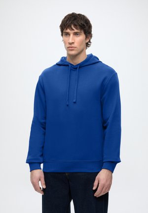 Homme aux cheveux bruns courts portant un sweat à capuche bleu et un pantalon foncé, debout devant un fond gris clair uni.