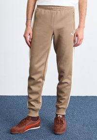Pantalones de chándal color beige con bolsillos laterales y rayas blancas, hechos de una tela suave. Combinados con zapatillas de deporte marrón oscuro con un acento rojo.
