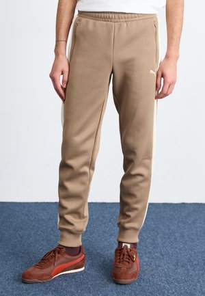 Pantalons de sport beige avec poches latérales et bandes blanches, fabriqués en tissu doux. Associés à des baskets marron foncé avec un accent rouge.