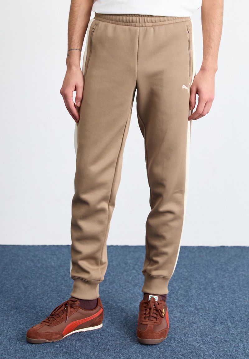 Pantalones de chándal color beige con bolsillos laterales y rayas blancas, hechos de una tela suave. Combinados con zapatillas de deporte marrón oscuro con un acento rojo.