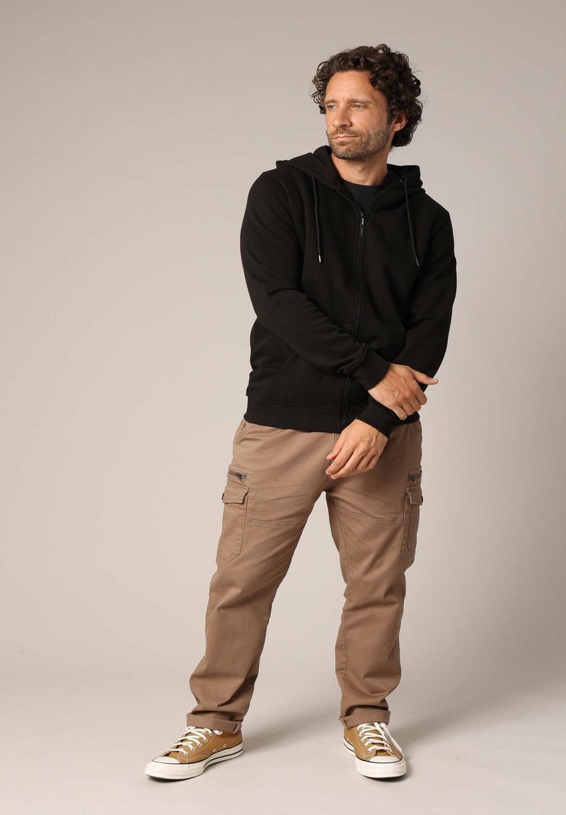 Zwarte zip-up hoodie met zijzakken, gecombineerd met beige cargobroek met pocketdetails en bruine sneakers met witte zolen.