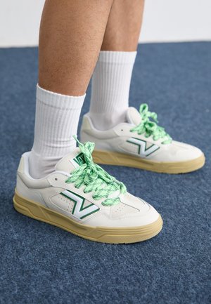 Valged tennisetüüpi saapad roheliste paelte ja detailidega, ümarate varvaste ning tekstuuriga tallaga. Kanda valgete lühikeste sokkidega sinisel pinnal.