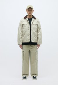 Μπεζ μπουφάν με καφέ κολάρο, μαύρο πουκάμισο, φαρδιά μπεζ cargo παντελόνια και μαύρα αθλητικά παπούτσια. Χαλαρή εμφάνιση με λειτουργικές τσέπες και απλό σχέδιο.