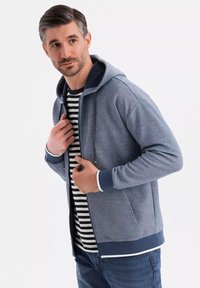 Hoodie zippé pour hommes en tissu bleu chiné, avec des finitions en bleu marine et des cordons de serrage, superposé sur un T-shirt rayé noir et blanc.