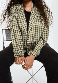 Personne aux longs cheveux bouclés assise sur une chaise en métal noir, portant un blazer vert à motifs sur une chemise noire et un pantalon noir, les mains jointes.