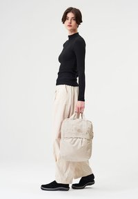 Beige Rucksack mit zwei oberen Griffen, gepolstertem Körper und zwei runden Akzenten. Kombiniert mit einem schwarzen Langarmshirt und hellen, lockeren Hosen.