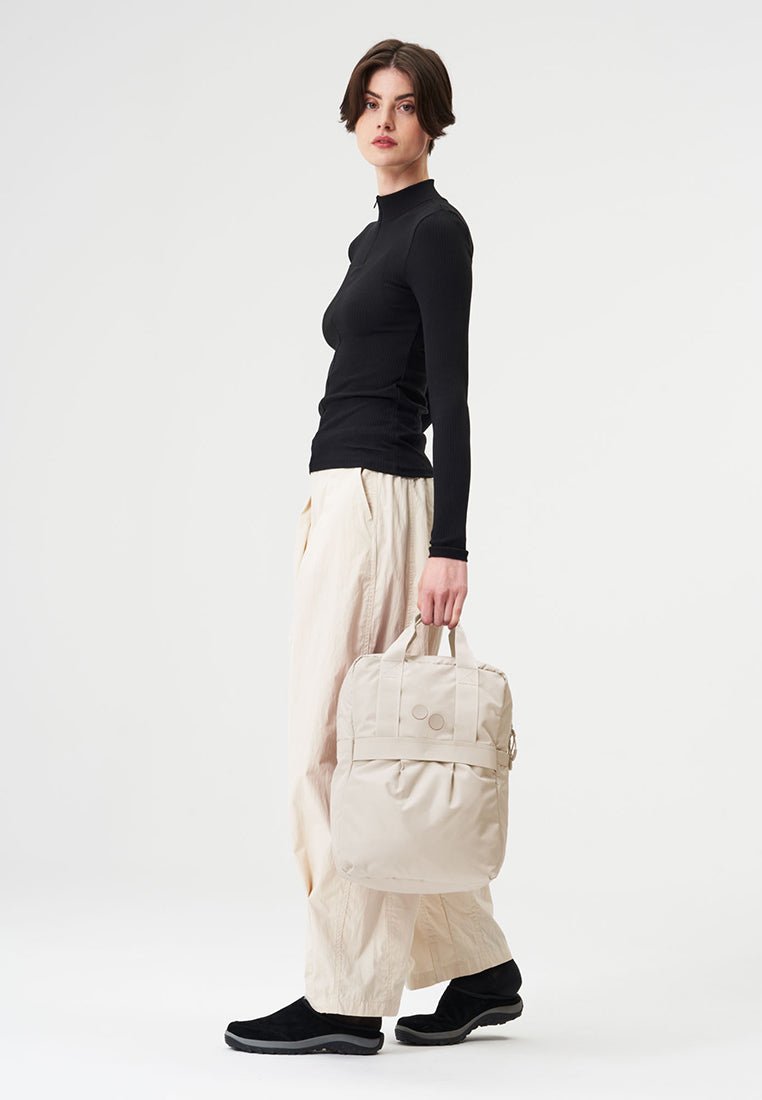 Beige Rucksack mit zwei oberen Griffen, gepolstertem Körper und zwei runden Akzenten. Kombiniert mit einem schwarzen Langarmshirt und hellen, lockeren Hosen.