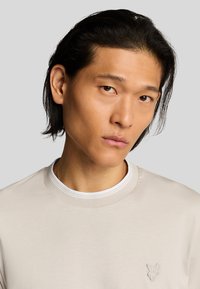 Helles beige T-Shirt mit kurzen Ärmeln und Rundhalsausschnitt, ausgestattet mit einem weißen Innenkragen und einem kleinen gestickten Schmetterlingslogo auf der Brust.