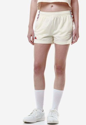 Pantaloni sportivi di colore-crema con vita elasticizzata, piping laterale blu e rosso, e dettaglio del logo davanti. Abbinati a calze e scarpe bianche.