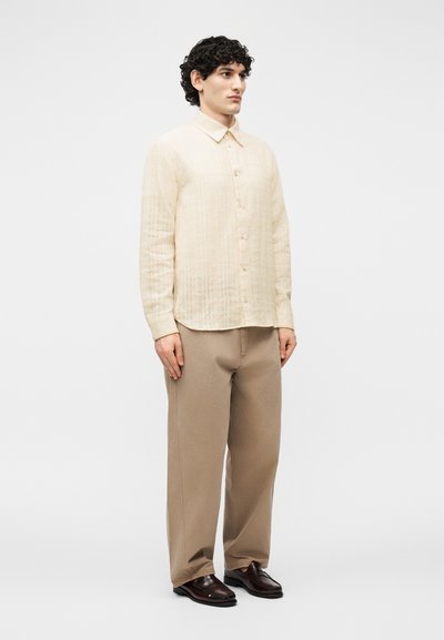 Άνδρας με σγουρά μαλλιά που φοράει μια εκρού πουκάμισο με μακριά μανίκια και κουμπιά, μπεζ φαρδιά παντελόνια και σκούρα καφέ μοκασίνια, στέκεται μπροστά σε απλό φόντο.