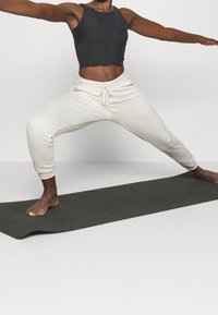Top cropped nero e pantaloni da felpa color crema, su un tappetino yoga scuro; i pantaloni hanno un laccio e polsini alle caviglie.