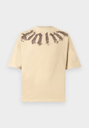 Tricou bej cu mânecă scurtă, croială oversized și design de text gotic de culoare maro închis, uzat, pe pieptul superior și umeri.
