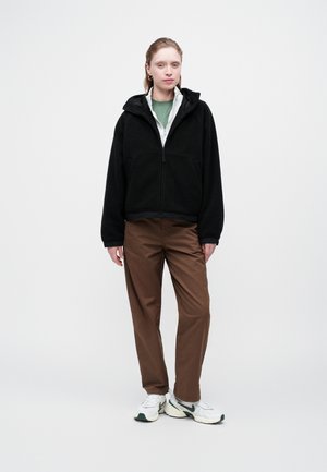 Femeie în picioare, purtând o jachetă neagră din fleece, o cămașă verde, pantaloni maro și adidași albi, pe un fundal simplu deschis.