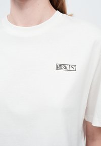 Cămașă albă cu text negru "ROSIE" și logo Puma pe partea stângă sus a pieptului, purtată de o persoană cu ten deschis și păr blond.