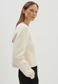 Cremefarbener Cardigan aus weichem Strickstoff. Merkmale sind ein gerippter Saum und Bündchen, eine durchgehende Knopfleiste und eine einzelne aufgesetzte Tasche auf der linken Seite.