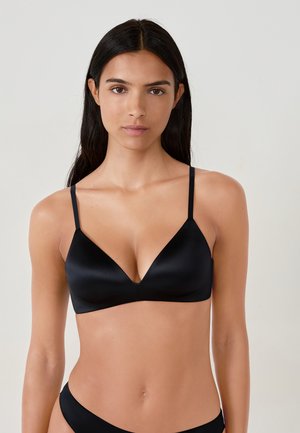 Donna con lunghi capelli scuri che indossa un bralette triangolare di satin nero e un bikini coordinato su uno sfondo chiaro e semplice.