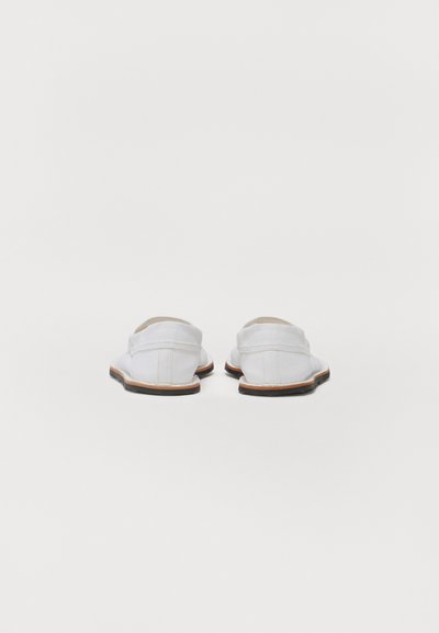 Hvide slip-on sko med en tekstiloverdel, rund tå og flad gummisål. Har syninger og en kontrasterende brun sålkant.