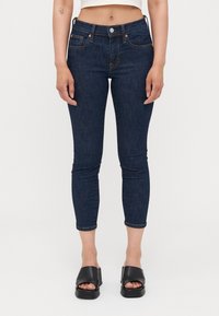 Jean skinny bleu foncé en denim, avec une taille mi-haute, un design à cinq poches et des coutures contrastantes. Associé à des sandales compensées noires.