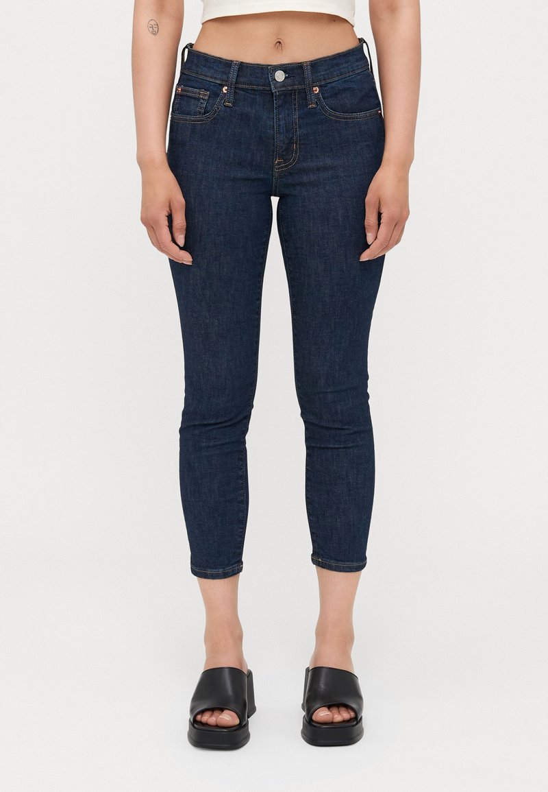 Jean skinny bleu foncé en denim, avec une taille mi-haute, un design à cinq poches et des coutures contrastantes. Associé à des sandales compensées noires.