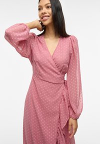 Abito avvolgente rosa realizzato in tessuto trasparente con maniche lunghe a palloncino, texture a pois e un orlo arricciato che accentua la vita.