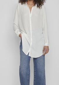 Camicetta bianca a maniche lunghe con un tessuto leggermente traslucido, orlo arrotondato e colletto classico, abbinata a jeans in denim blu a gamba larga.