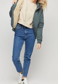 Gepolsterte grüne Bomberjacke mit einem beigen Kunstpelzfutter, kombiniert mit hoch taillierten blauen Jeans und weißen Sneakers mit grünen Akzenten.