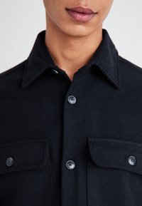 Chemise noire à manches longues avec un col structuré, deux poches poitrine et des boutons noirs ; fabriquée à partir d'un matériau durable avec une texture lisse.