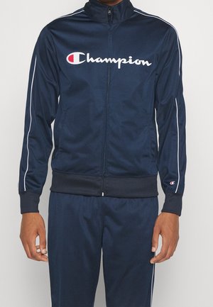 Hombre con chaqueta de chándal Champion azul marino y pantalones a juego con detalles de piping blancos en las mangas y las piernas.
