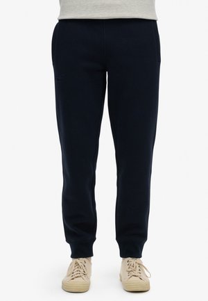 Marineblå sweatpants lavet af blødt stof, med en tapered pasform, to sidelommer og elastiske manchetter, parret med beige sneakers.