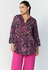 Mehrfarbige, gemusterte Bluse mit V-Ausschnitt und Dreiviertelärmel, ergänzt durch pinkfarbene, weite Hosen. Bemerkenswerte Details umfassen ein geometrisches Design.
