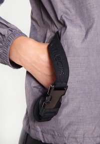 Černý nastavitelný pásek s kovovou sponou připevněný k šedé texturované bundě s nařasenými manžetami, na fotografii je vidět ruka uchopující pásek.