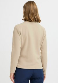 Beige fleece-takki pehmeällä tekstuurilla, pitkät hihat ja suora leikkaus. Saumaton muotoilu, jossa on minimaalinen varustus ja ei voimakkaita yksityiskohtia.