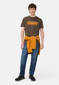 Brun t-shirt med "CHASE NEW HORIZONS" grafik i orange, parret med blå denim jeans og en tekstureret orange sweater bundet omkring livet. Sorte sko.