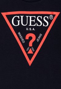 Marineblå bomull T-skjorte med en stor rød triangel-logo med teksten "GUESS" og "U.S.A.", et sentralt spørsmåletegn og detaljer om "WASHED JEANS".
