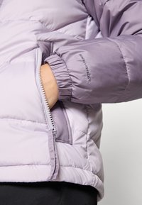 Purpurinė pūkinė striukė su užtrauktuku ir elastingomis rankovėmis. Medžiaga yra lygaus paviršiaus su blizgiu tekstūru.