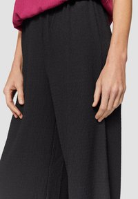 Zwarte, gestructureerde wijde broek met een elastische tailleband. Een hand is nabij de taille gepositioneerd, waardoor de gekreukte uitstraling van de stof wordt benadrukt.