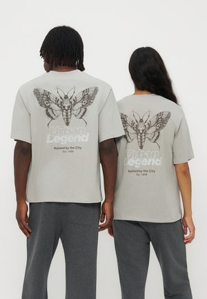 To personer står side om side iført lysegrå T-shirts med en stor detaljeret natsommerfugl og "Urban Legend Raised by the City" tekst på ryggen.