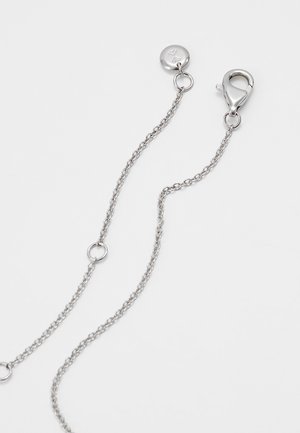 Collier chaîne en argent avec fermoir homard et petite étiquette ronde reliés par de fines mailles sur fond blanc.