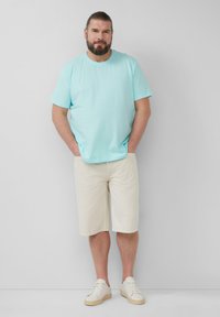 Lichtblauwe katoenen t-shirt, beige shorts tot op de knie, witte sneakers, casual fit, ontspannen stijl, man staat met zijn handen in zijn zakken.