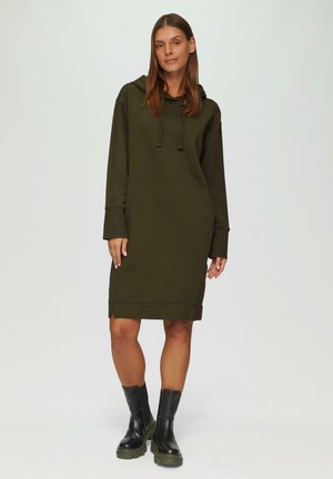 Olivegrünes Kapuzenkleid aus weichem Stoff, mit langen Ärmeln, Seitenschlitzen am Saum und kombiniert mit schwarzen, chunky Knöchelschuhen.