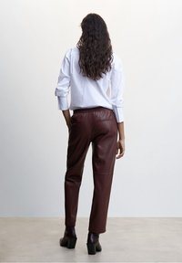 Femme aux longs cheveux ondulés, de dos, portant une chemise blanche, un pantalon en cuir marron foncé et des bottines à talons sur un fond uni.