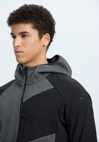 Giacca con cappuccio caratterizzata da una combinazione di materiali testurizzati in nero e grigio, con chiusura a zip e dettaglio di tasca sul petto con zip.