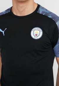 Musta urheilupaita lyhyillä hihoilla, jossa on sininen kuviollinen olkakuvio. Edessä on pyöreä Manchester City -logo.