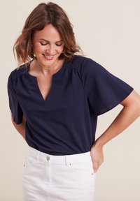 Maglietta blu navy a maniche corte con scollatura a V, in tessuto morbido e con un leggero arriccio sulle spalle, abbinata a jeans bianchi.