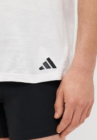 Weißes T-Shirt mit schwarzem Adidas-Logo-Akzent an der unteren linken Seite. Kurze Ärmel, Rundhalsausschnitt, aus weichem Baumwollmaterial. Schwarze Shorts sind sichtbar.