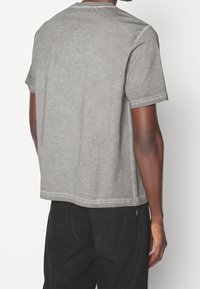 Szara bawełniana koszulka z krótkim rękawem, okrągłym dekoltem i bocznymi rozcięciami. Posiada widoczne przeszycia wzdłuż szwów oraz luźny krój.