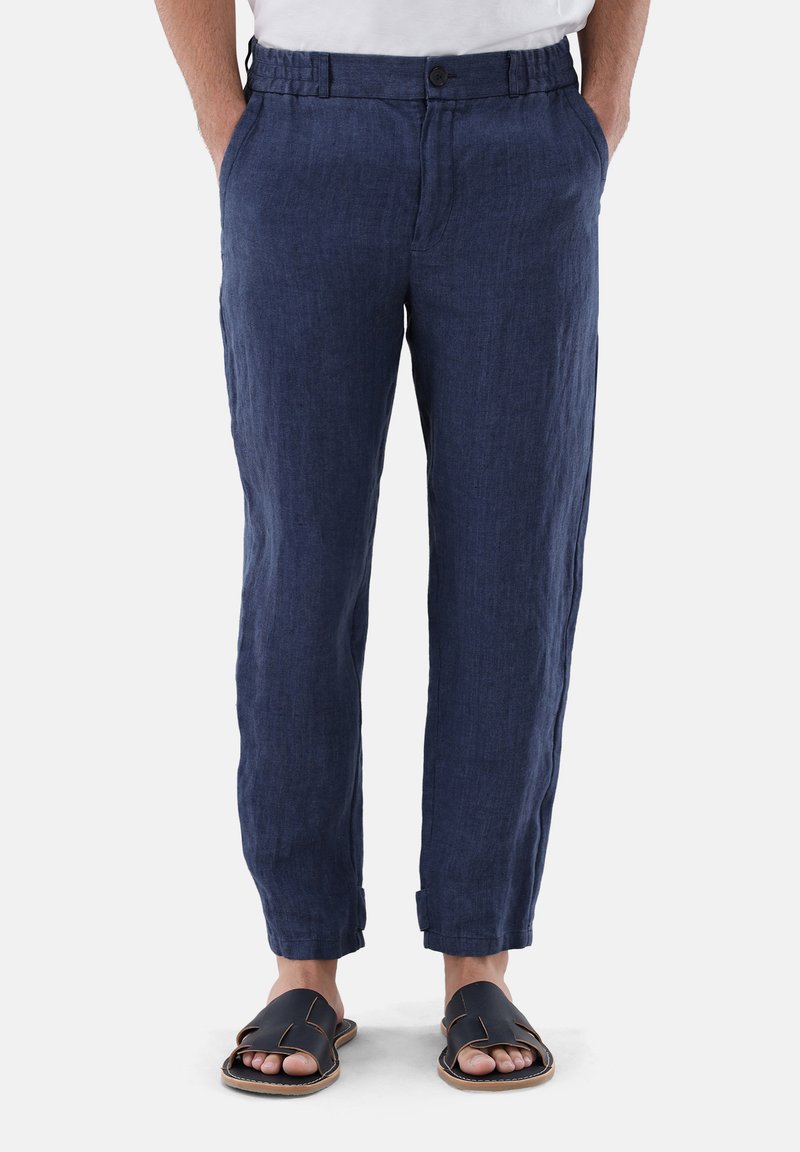 Marineblaue Leinenhose mit lässiger Passform, Paspeldetails und hochgerollten Säumen. Kombiniert mit schwarzen flachen Sandalen.
